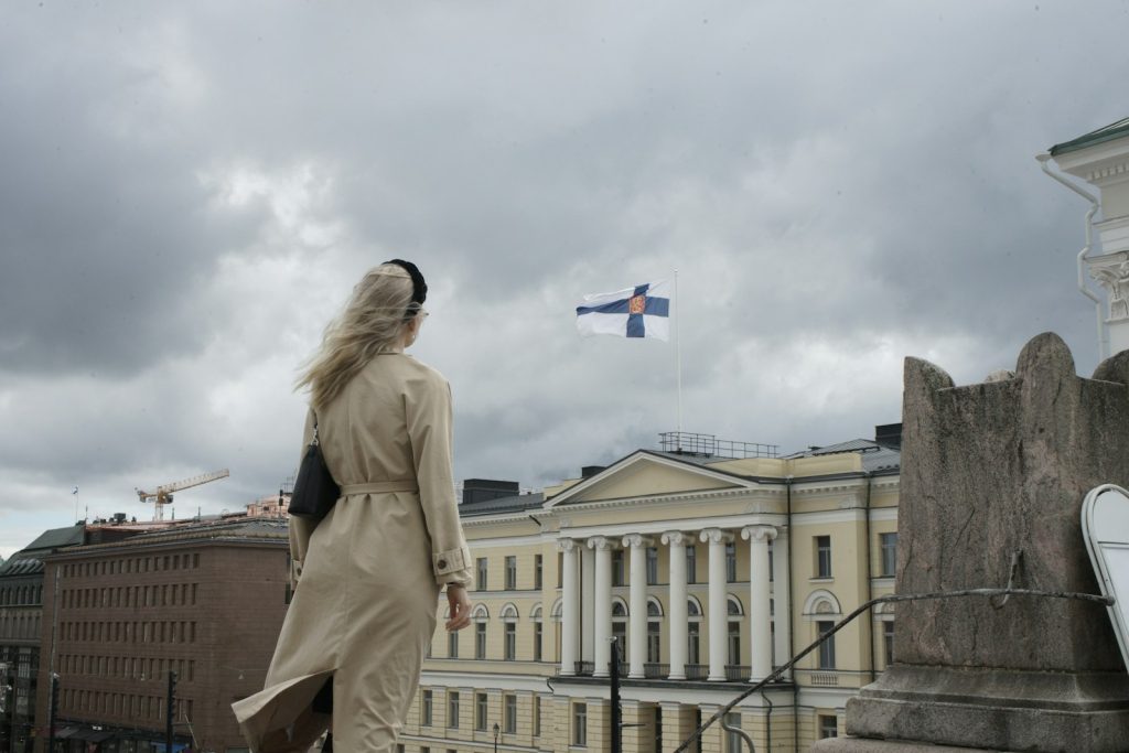 A woman in a beige trench coat on a city street under a cloudy sky