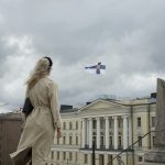 A woman in a beige trench coat on a city street under a cloudy sky