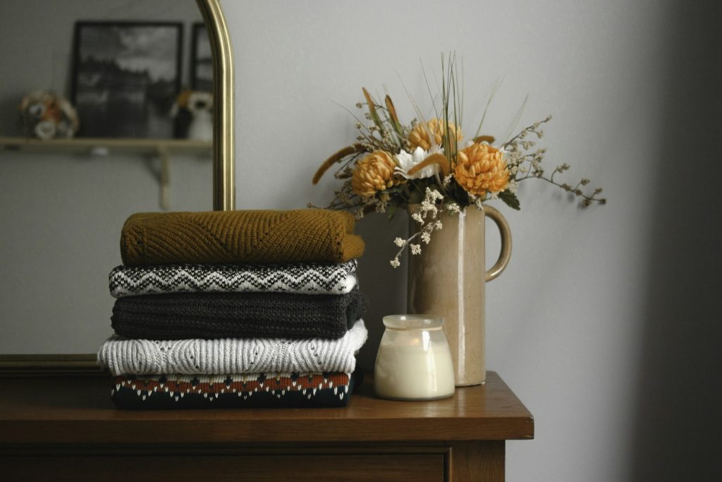 Folded knitwear stacked on a wooden dresser beside dried flowers