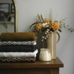 Folded knitwear stacked on a wooden dresser beside dried flowers